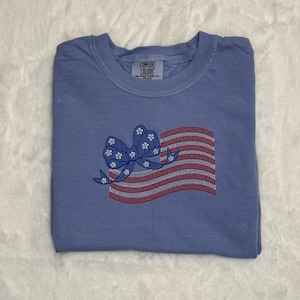 May include: A blue t-shirt with an embroidered American flag design. The flag features red and white stripes and a blue bow with white flower accents. The shirt is folded and has a Comfort Colors tag.