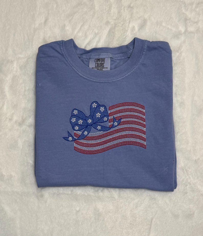 May include: A blue t-shirt with an embroidered American flag design. The flag features red and white stripes and a blue bow with white flower accents. The shirt is folded and has a Comfort Colors tag.