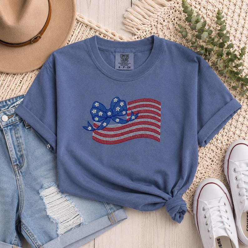 May include: A blue Comfort Colors t-shirt featuring an embroidered American flag design with a bow. The flag has red and white stripes, and the bow is blue with white flower accents. The shirt is paired with denim shorts and white sneakers.