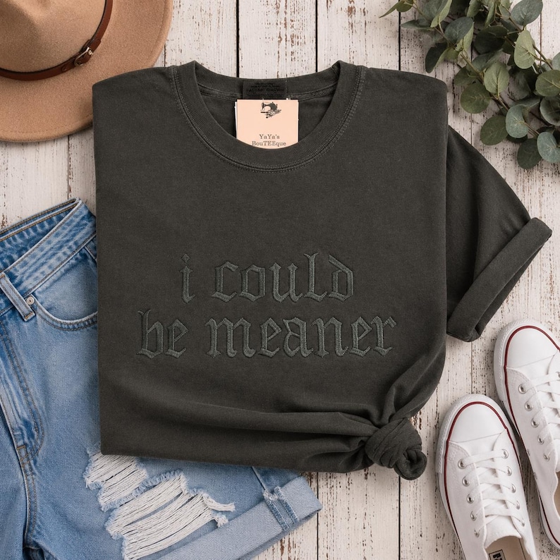 Puede incluir: Camiseta gris oscuro con la frase bordada "I COULD be MEANER." La camiseta est&aacute; doblada, con una etiqueta "YaYa's Boutique." Se combina con vaqueros azules, un sombrero marr&oacute;n y zapatillas blancas.