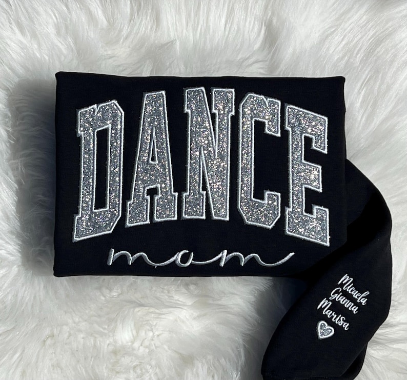May include: Black sweatshirt with silver glitter lettering that reads "DANCE mom". The sleeve has white lettering that reads "Michaela, Gianna, Marisa" with a heart symbol.