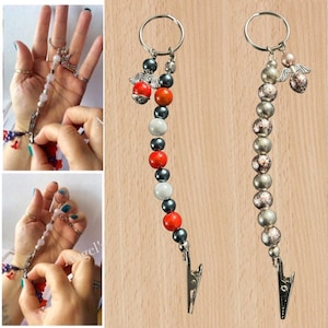 May include: Two beaded keychains with metal clips. One has red, white, and dark grey beads, a silver angel charm and a red bead. The other has silver and patterned beads, with a silver angel charm.