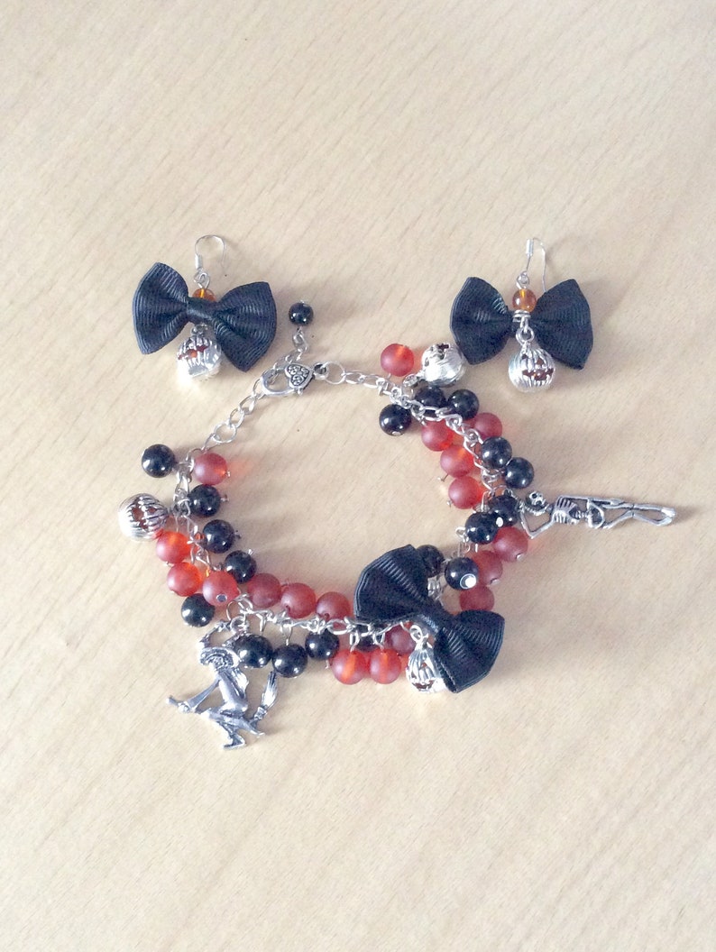 May include: A Halloween-themed charm bracelet with red and black beads, a black bow, and various charms, including a pumpkin, a skeleton, and a witch. The bracelet also includes a pair of matching earrings with black bows and pumpkin charms.