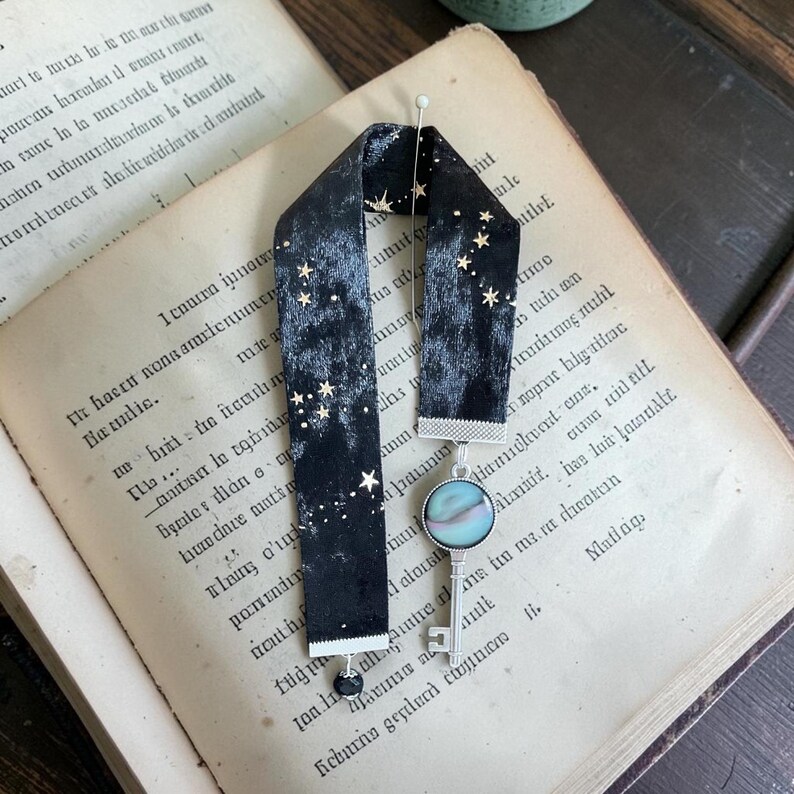 May include: Black velvet bookmark with gold star detailing and a silver key pendant. The bookmark is shown on an open book with aged, yellowed pages and handwritten text.