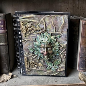 May include: A spiral-bound journal with a sculpted cover depicting a Green Man face encircled by leaves. The cover has a textured, earthy look with green, brown, and gold tones. The journal is on a wooden shelf with antique books.