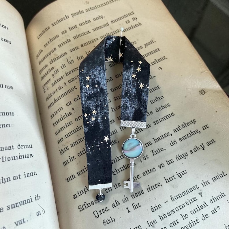 May include: A black velvet bookmark with gold star detailing and a key charm. The bookmark is placed in an open book with aged pages. The key charm features an iridescent stone and a silver-coloured key.