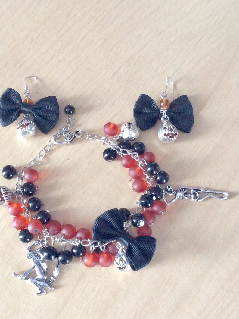 May include: A black and red beaded bracelet with charms, including a black bow, a skeleton, and a pumpkin. The bracelet also includes two matching earrings with black bows and pumpkin charms.