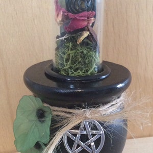 May include: Black cauldron with a pentagram charm, a green leaf, and a glass vial filled with dried flowers, moss, and a black spiral incense.
