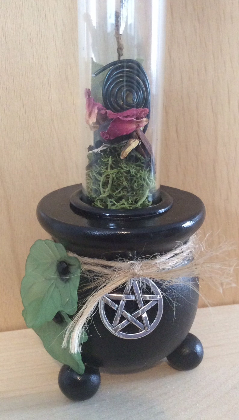 May include: Black cauldron with a pentagram charm, a green leaf, and a glass vial filled with dried flowers, moss, and a black spiral incense.