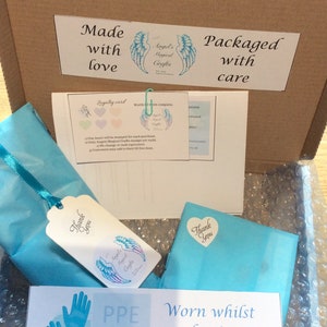 May include: A cardboard box with a blue and white tissue paper-wrapped package inside. The box is labeled "Made with love" and "Packaged with care." The tissue paper has a "Thank you" sticker with a heart on it. The box also has a sticker with a pair of blue gloves and a blue face mask, labeled "PPE Worn whilst packaging." The sticker also has the text "Stay alert - Control the virus - Save lives."