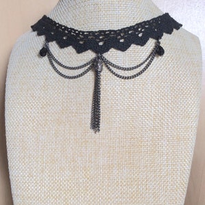 May include: A black lace choker necklace with a silver chain and tassel pendant. The necklace has two small silver beads on each side of the pendant.