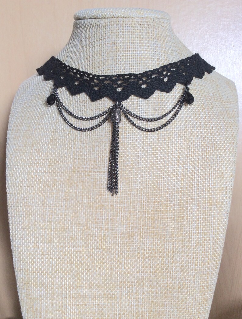 May include: A black lace choker necklace with a silver chain and tassel pendant. The necklace has two small silver beads on each side of the pendant.