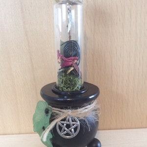 May include: A black cauldron with a pentagram on the front, holding a glass bottle filled with dried herbs, flowers, and a metal spiral. The bottle has a cork stopper and a silver charm hanging from it.