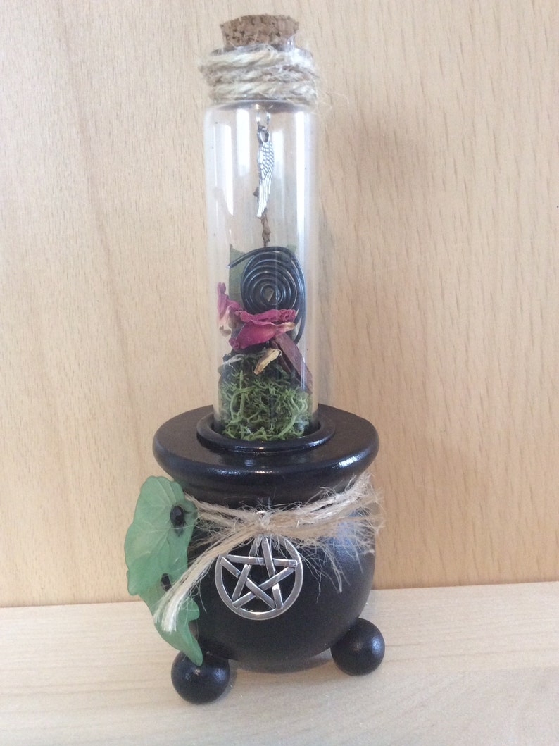 May include: A black cauldron with a pentagram on the front, holding a glass bottle filled with dried herbs, flowers, and a metal spiral. The bottle has a cork stopper and a silver charm hanging from it.