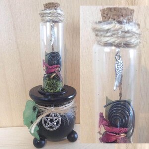 May include: A black cauldron with a pentagram on the front, holding a glass bottle with a cork stopper. The bottle contains dried rose petals, moss, a black licorice twist, and a silver charm with a wing design.