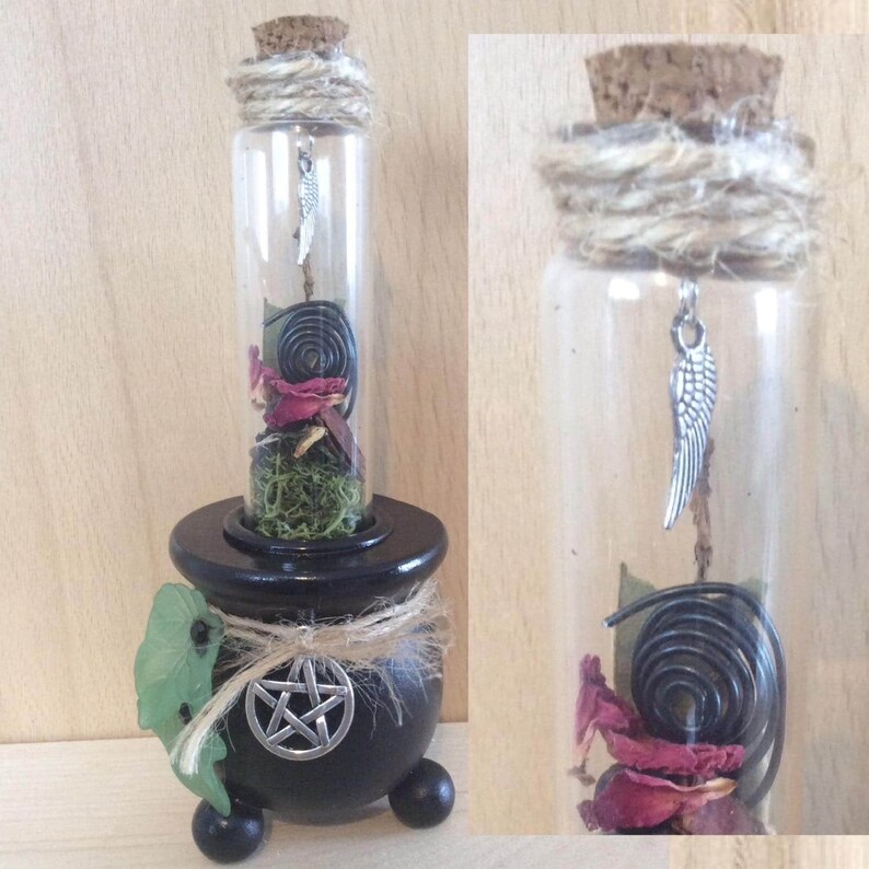 May include: A black cauldron with a pentagram on the front, holding a glass bottle with a cork stopper. The bottle contains dried rose petals, moss, a black licorice twist, and a silver charm with a wing design.