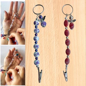 May include: Two beaded keychain accessories. One features purple and white floral beads, the other has red and white oval beads. Both have a silver-tone clip and angel charm, attached to a keyring.