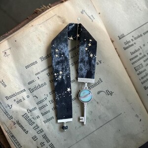 May include: A black velvet bookmark with gold star detailing and a silver key charm featuring a pearlescent stone. The bookmark is shown on an open book with aged pages.