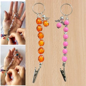 May include: Two beaded keychains with silver clips. One has orange, textured beads and a small angel charm. The other has pink beads and a silver heart charm. Both have a silver ring at the top. The beads are approximately 1 cm in diameter.