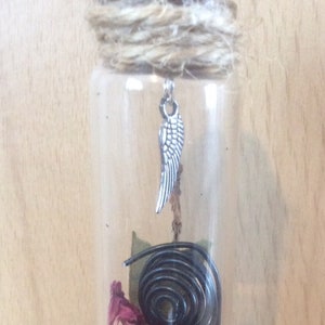 May include: A glass bottle with a cork top and a silver angel wing charm. The bottle contains dried rose petals, a black licorice twist, and a green leaf.