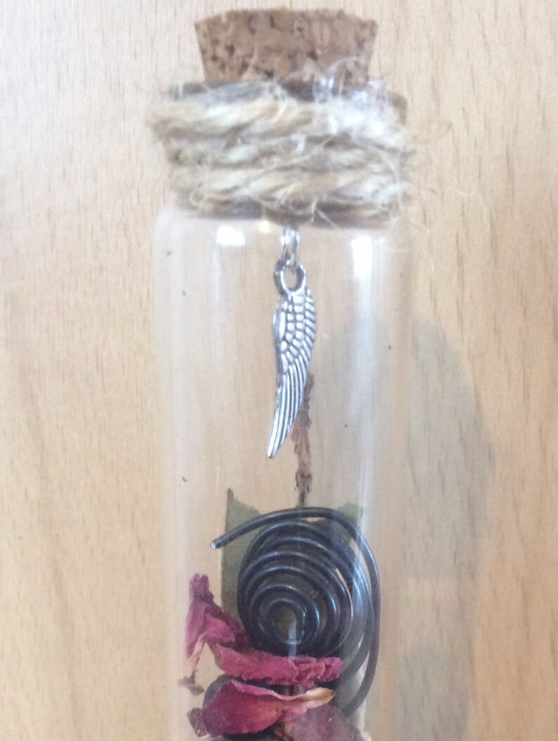 May include: A glass bottle with a cork top and a silver angel wing charm. The bottle contains dried rose petals, a black licorice twist, and a green leaf.