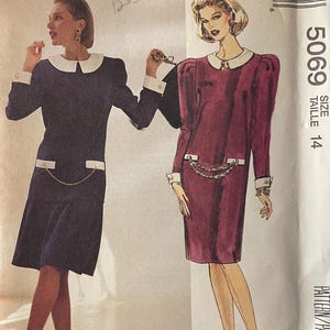 May include: McCall's sewing pattern 5069 featuring two dress designs. The dresses have a white collar and cuffs, with a decorative chain detail at the waist. One dress is navy blue, the other is burgundy. The pattern includes the text "LIZ ROBERTS".