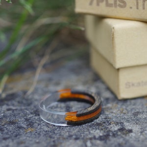 May include: A clear resin ring with black and orange wood inlay. The ring is sitting on a grey surface next to a stack of boxes. The top box has the text "7PLIS.fr" printed on it.