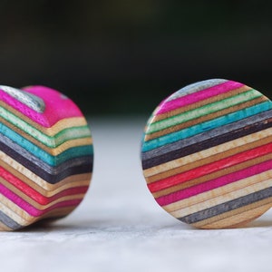 Könnte beinhalten: Zwei Holz-Ohrstecker mit farbenfrohem Streifendesign. Einer ist herzförmig, der andere rund. Die Ohrstecker bestehen aus geschichtetem Holz in Rosa-, Blau-, Grün- und Brauntönen.