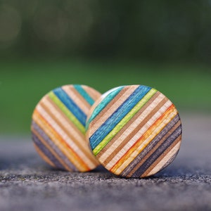 Peut inclure: Deux jauges d'oreille rondes, colorées et rayées en bois, fabriquées à partir de plateaux de skate recyclés. Les jauges d'oreille sont sur une surface brune. Le texte "7PLIS Skateboard recycle" est visible sur une boîte brune en arrière-plan.