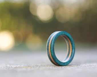 May include: A green and brown wooden ring with a thin, light brown stripe. The ring is lying on a gray surface.
