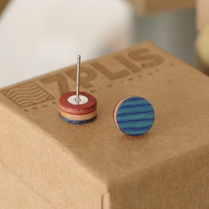 Puede incluir: Un par de pendientes de madera, uno con un diseño de rayas rojas y blancas y el otro con un diseño de rayas azules y blancas. Los pendientes están sobre una caja marrón con el texto "7PLUS" impreso en ella.