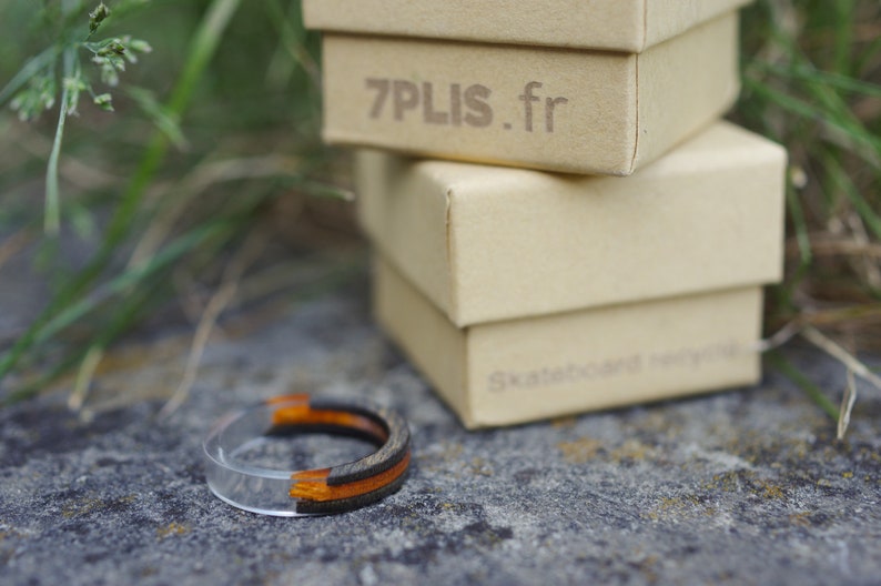 May include: A clear resin ring with black and orange wood inlay. The ring is sitting on a grey surface next to a stack of boxes. The top box has the text "7PLIS.fr" printed on it.