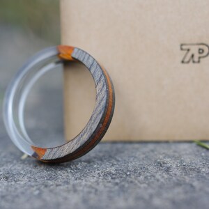 May include: A wooden ring with a silver band and a brown and orange wood inlay. The ring is resting on a grey surface next to a brown box with the letters "TP" printed on it.