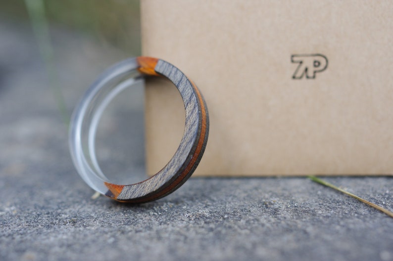 May include: A wooden ring with a silver band and a brown and orange wood inlay. The ring is resting on a grey surface next to a brown box with the letters "TP" printed on it.