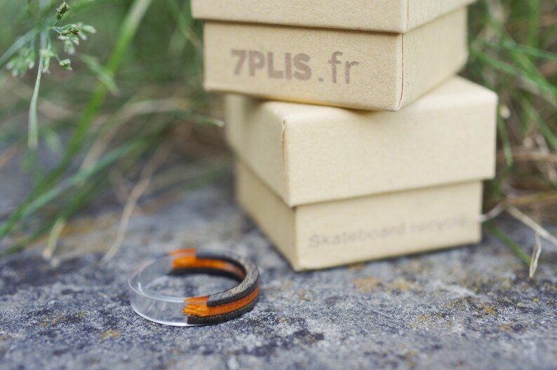 May include: A clear resin ring with black and orange wood inlay. The ring is sitting on a grey surface in front of three boxes. The top box has the text "7PLIS.fr" printed on it.