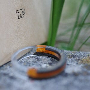 May include: A clear resin ring with a black and orange wood inlay. The ring is sitting on a grey stone surface.