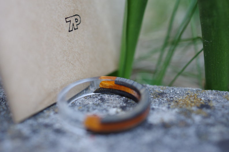 May include: A clear resin ring with a black and orange wood inlay. The ring is sitting on a grey stone surface.