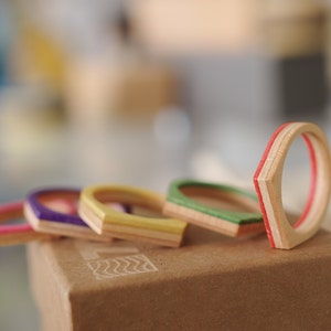 May include: Five wooden rings with different coloured stripes. The rings are stacked on top of each other and are sitting on a brown box.