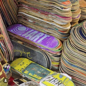Op de afbeelding: Een stapel gebruikte skateboarddecks, sommige met graphics, waaronder een paarse deck met het woord "JART" erop gedrukt.