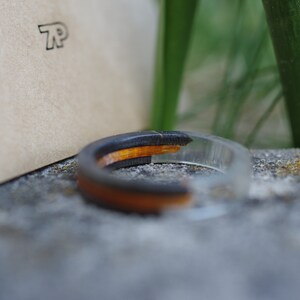 May include: A black and orange resin ring with a clear resin band. The ring is sitting on a grey stone surface.