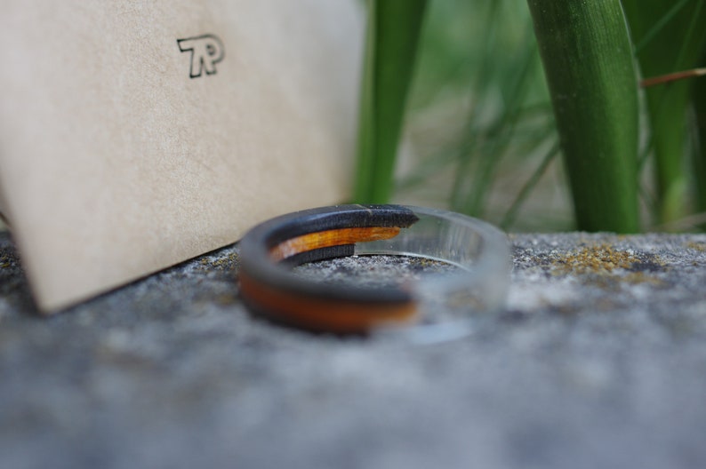 May include: A black and orange resin ring with a clear resin band. The ring is sitting on a grey stone surface.