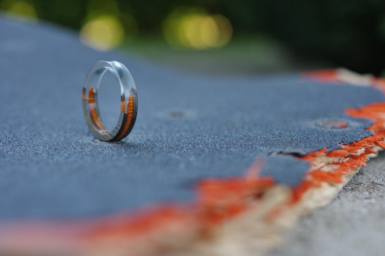 May include: A clear resin ring with a thin band of wood inlay. The ring is sitting on a grey and orange surface.