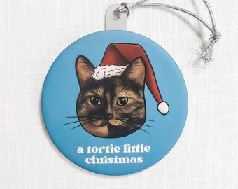 May include: Round Christmas ornament with a light blue background featuring a cat wearing a Santa hat. The cat has a tortie pattern and the text "a tortie little christmas" is printed below the cat's face. A silver string is attached for hanging.