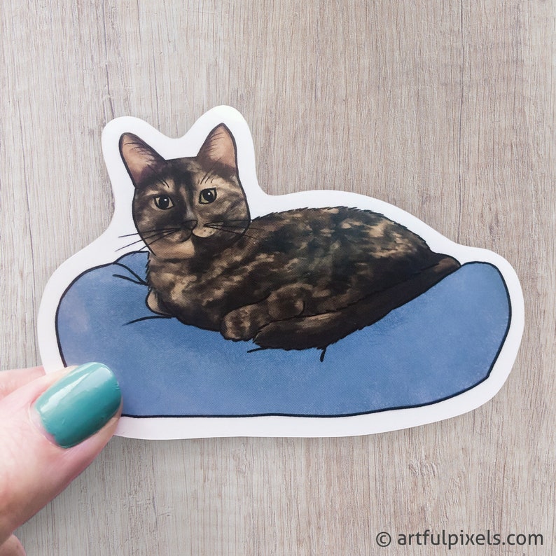 May include: A sticker of a brown tabby cat with green eyes lying on a blue pillow. The cat is looking directly at the viewer.