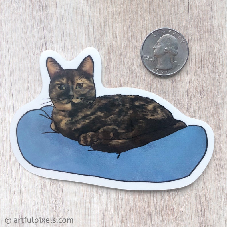 May include: A sticker of a brown tabby cat with green eyes lying on a blue pillow. The cat is looking directly at the viewer.
