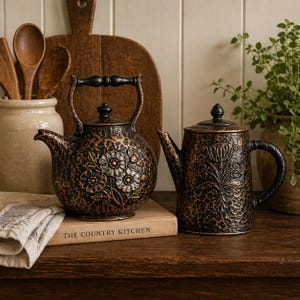 May include: Two decorative teapots with a textured, bronze-colored finish. One is round with floral detailing, the other is rectangular with a floral design. The teapots sit on a wooden surface next to a book that reads "THE COUNTRY KITCHEN".