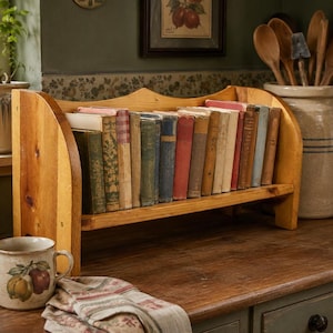 May include: A wooden desktop bookshelf with arched side panels holding a row of vintage hardcover books with various colored spines. The shelf is constructed from light-toned wood with a visible grain, featuring a simple open design for storage.