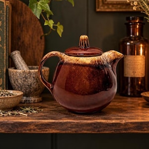 May include: A round ceramic teapot featuring a glossy dark brown glaze that transitions into a lighter cream-colored band near the top. The teapot has a curved handle, a short spout, and a matching lid with a small, ribbed, vertical handle.