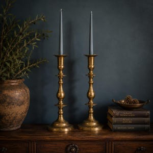 May include: Two tall, polished brass candlesticks with slender, tapered stems and wide circular bases, each holding a single slim, dark grey taper candle. The candlesticks feature a classic turned design with multiple decorative rings.