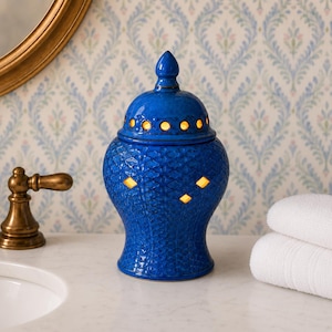 May include: A cobalt blue ceramic jar with a lid, featuring a textured scale pattern and cut-out diamond and circular shapes that emit a warm internal glow. The vessel has a rounded body, a tapered neck, and a finial top, standing upright.